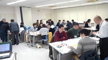 明るい室内でグループごとに机を分けて話し合う人たちの写真