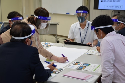 白い机を囲んで紙に書き込んでいく6人の男女の写真