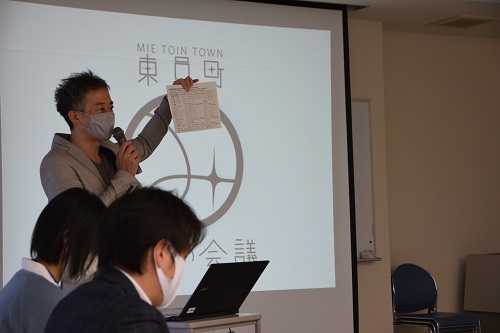 スクリーンを背に資料を示しながら話す男性と俯きながら聞いている2人の写真