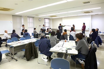 ホワイトボードの前で紙を広げる二人の人とそれを見る大勢の人達の写真