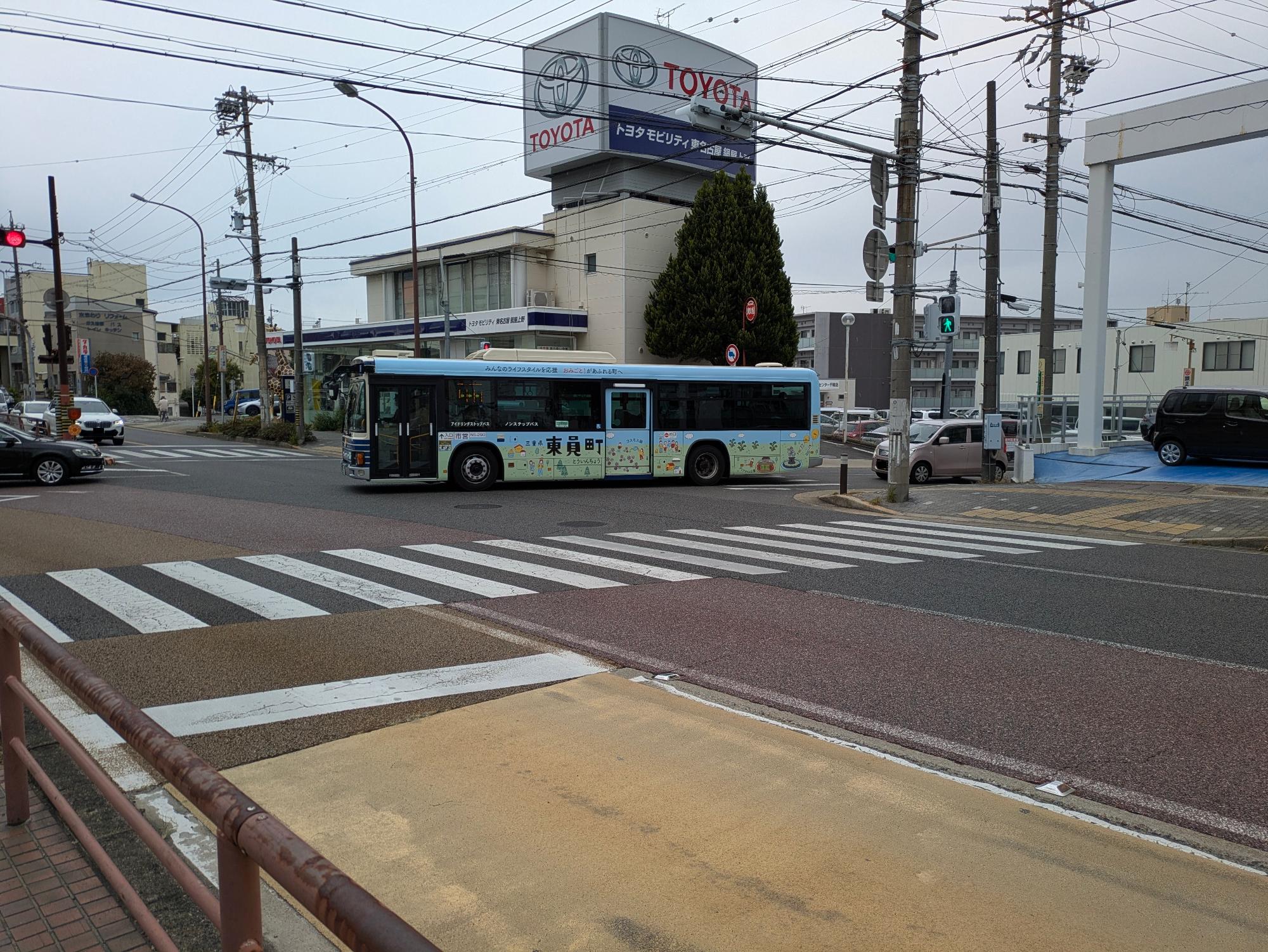 名古屋市大森営業所系統を走行する水色を基調とした東員町ポスターデザインにラッピングした名古屋市バスが交差点を右折する様子