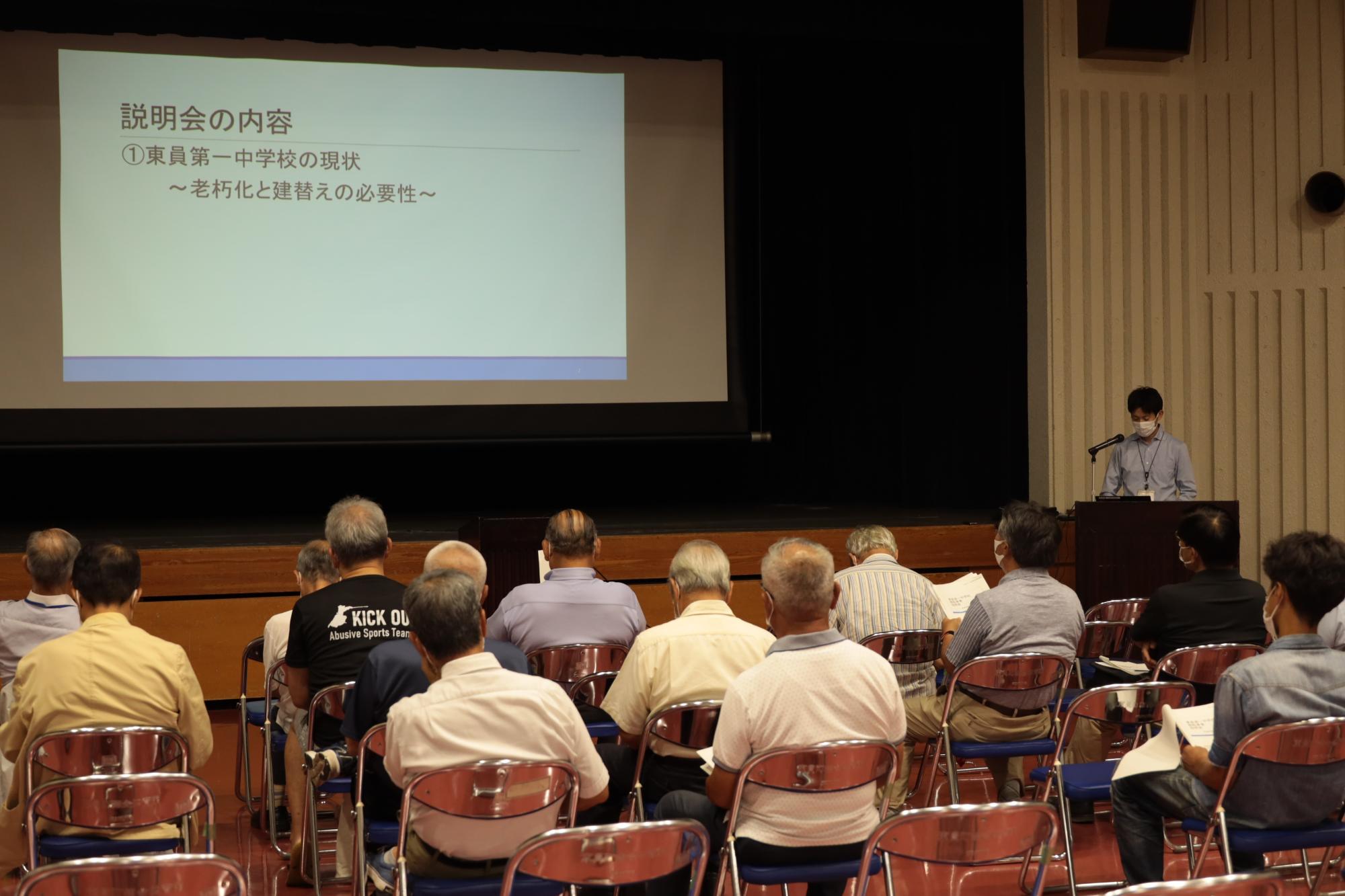 説明会の内容を司会が進行し町民たちがスクリーンを見ながら聞いている写真