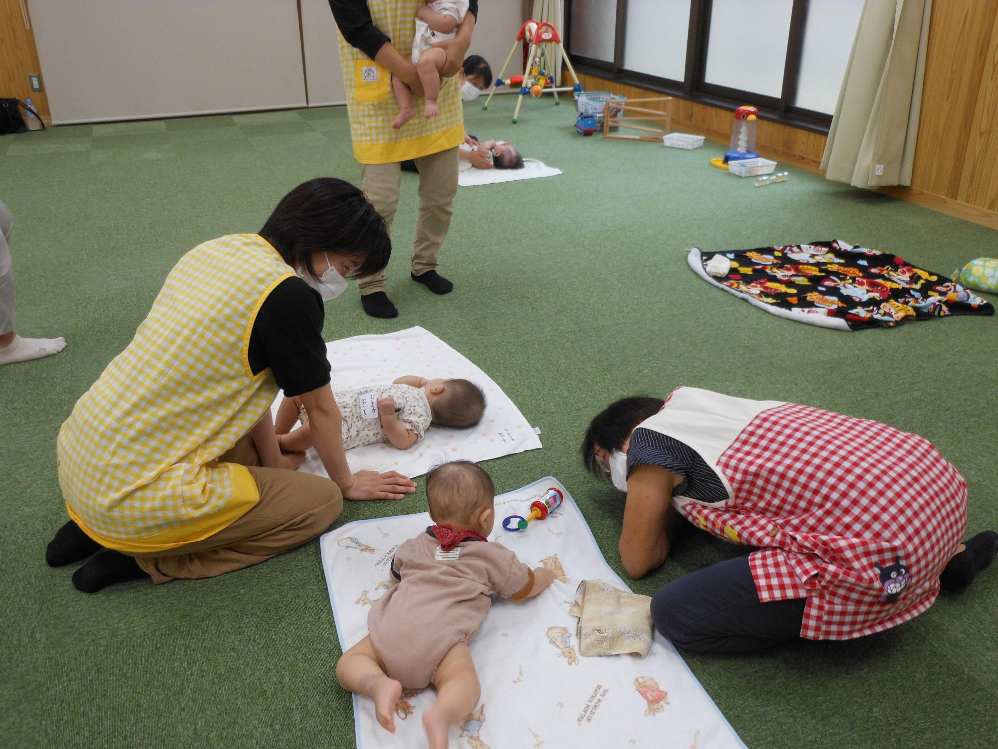 タオルの上に寝転がる赤ちゃんと相手をする女性たちの写真