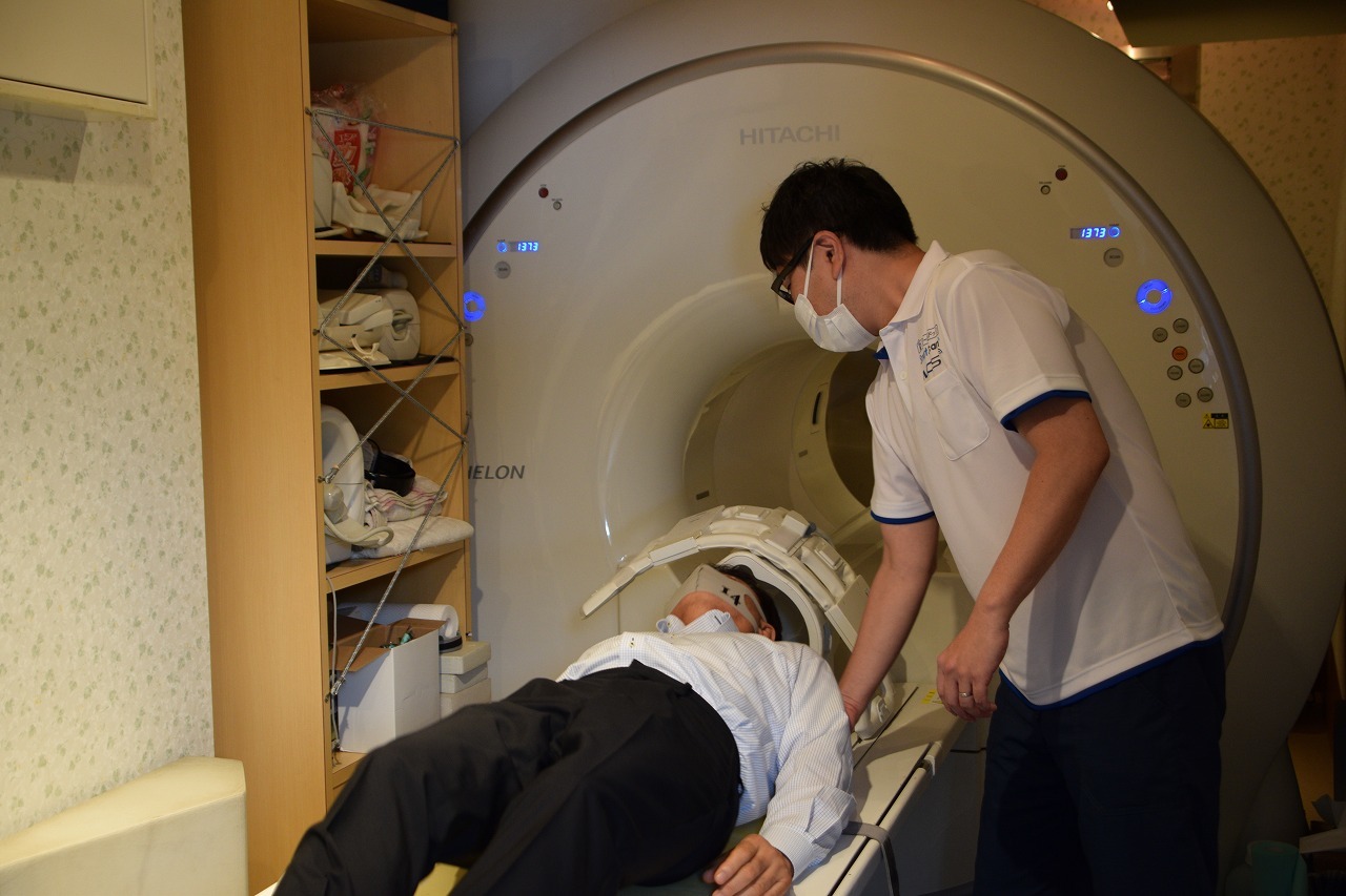 MRIのベッドに横になる男性と介助する白衣を着てマスクをした人の写真