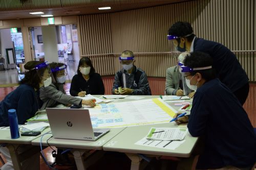 室内で、机の中央に大きな紙を広げて話し合っている人たちの写真