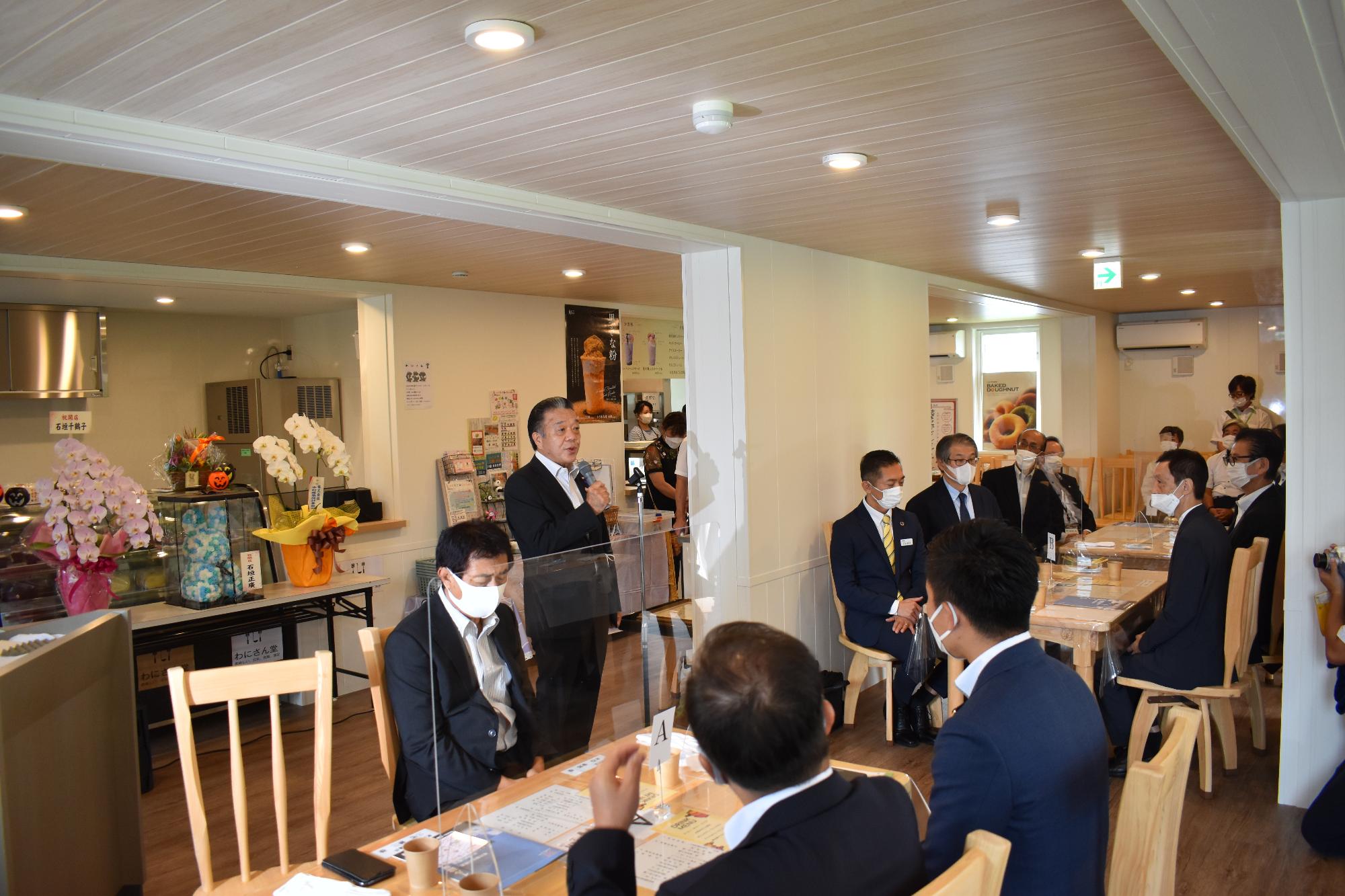 マメマチCAFEの中で挨拶をする三宅議長とそれをテーブルで聞く参加者たちの写真